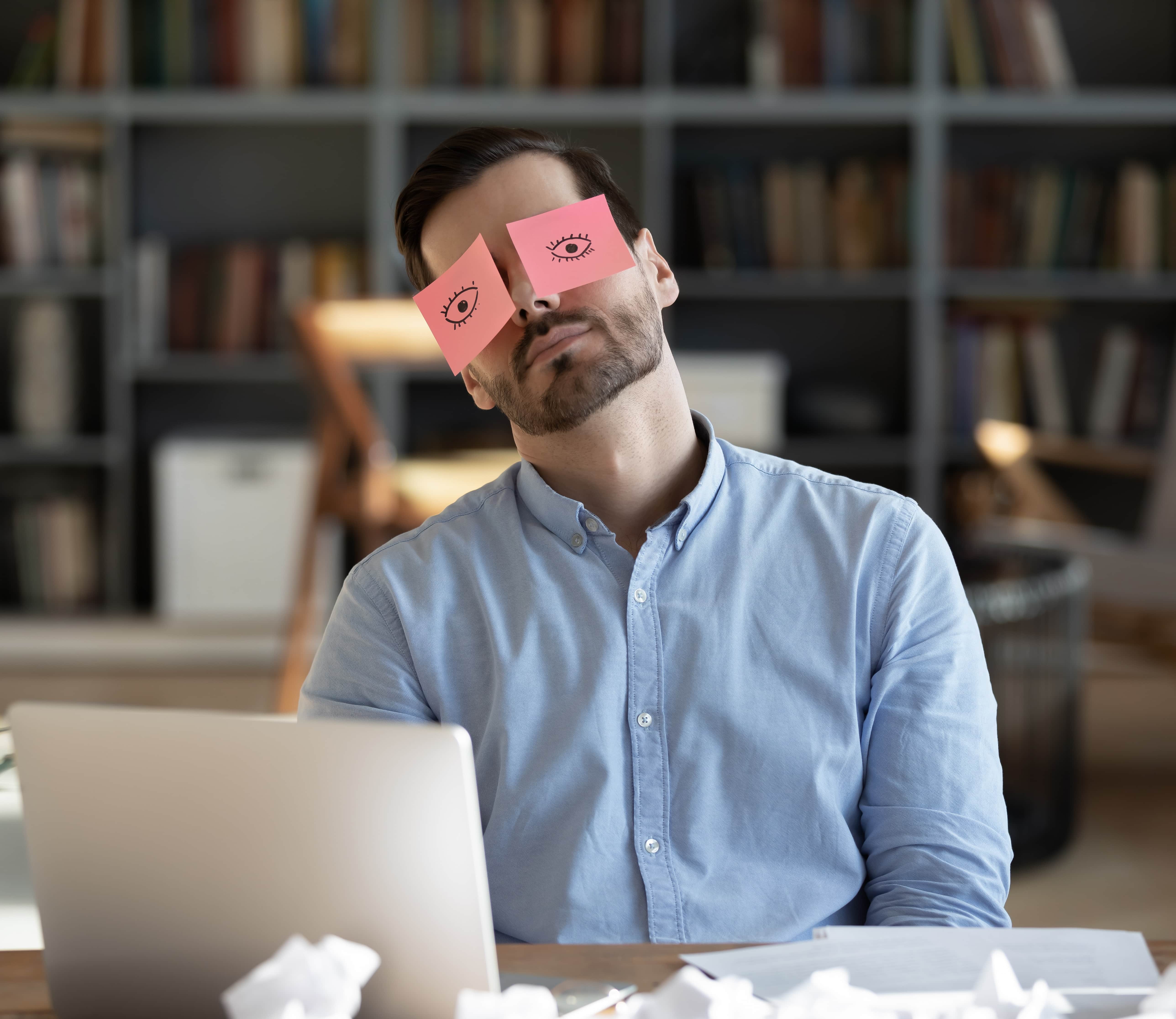 Man at a desk with two pink sticky notes covering his eyes, each with a drawn eye, in a blurred bookshelf-filled room.