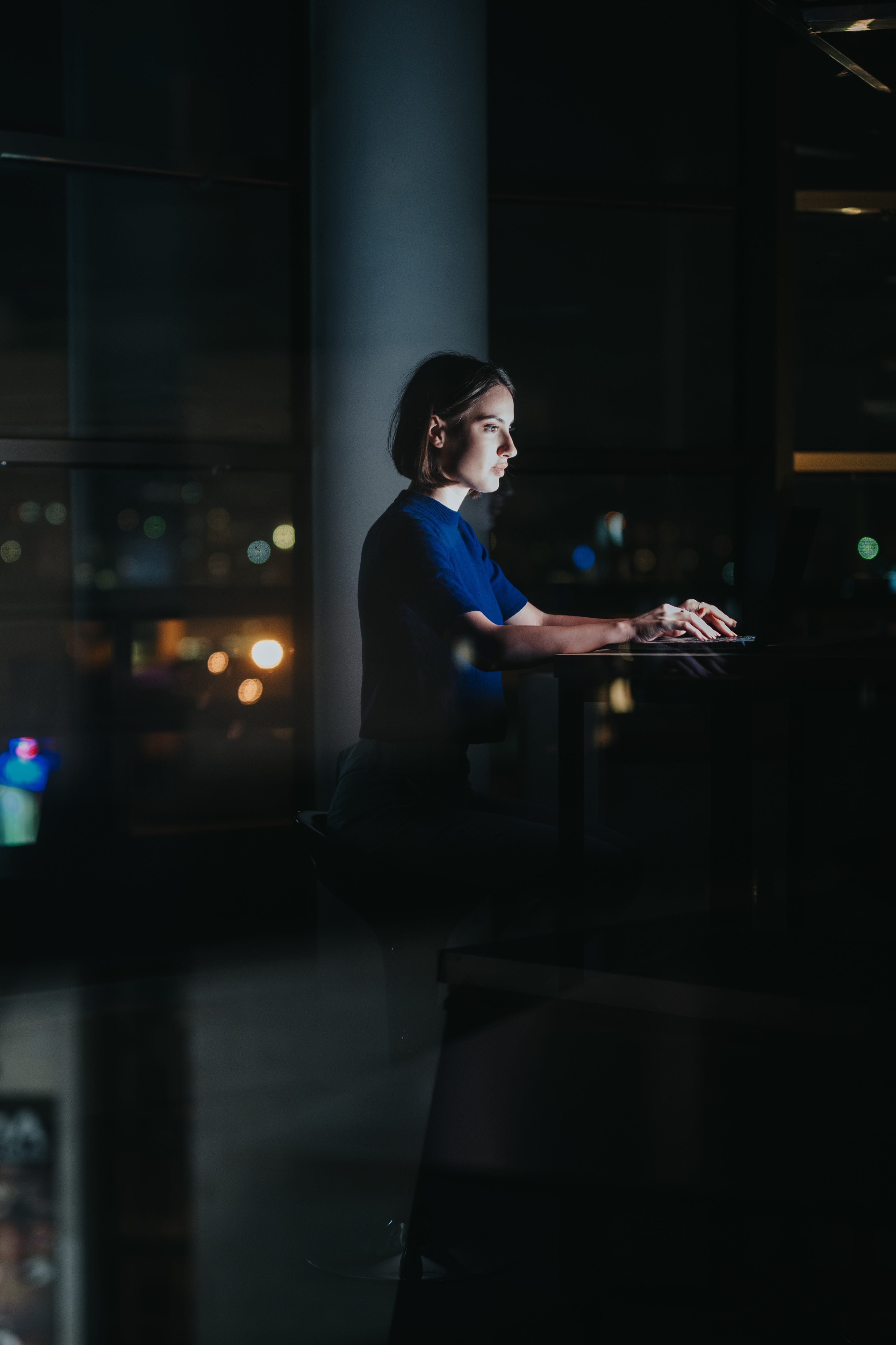 Woman sitting at a high desk, lit by a laptop screen, working in a dark room with city lights outside the window