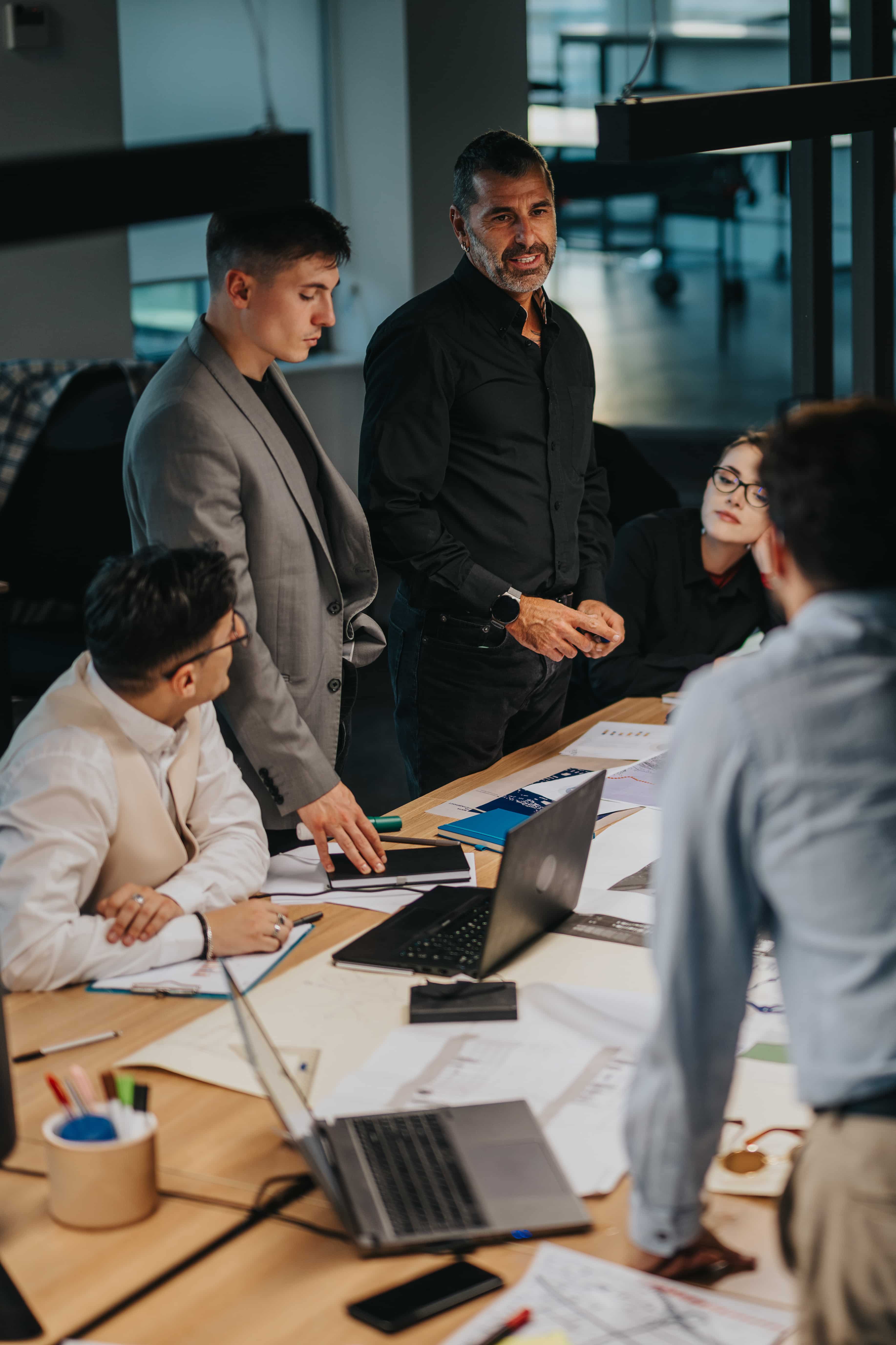 Team of professionals standing and seated around a table with laptops and documents during a meeting in a modern office.