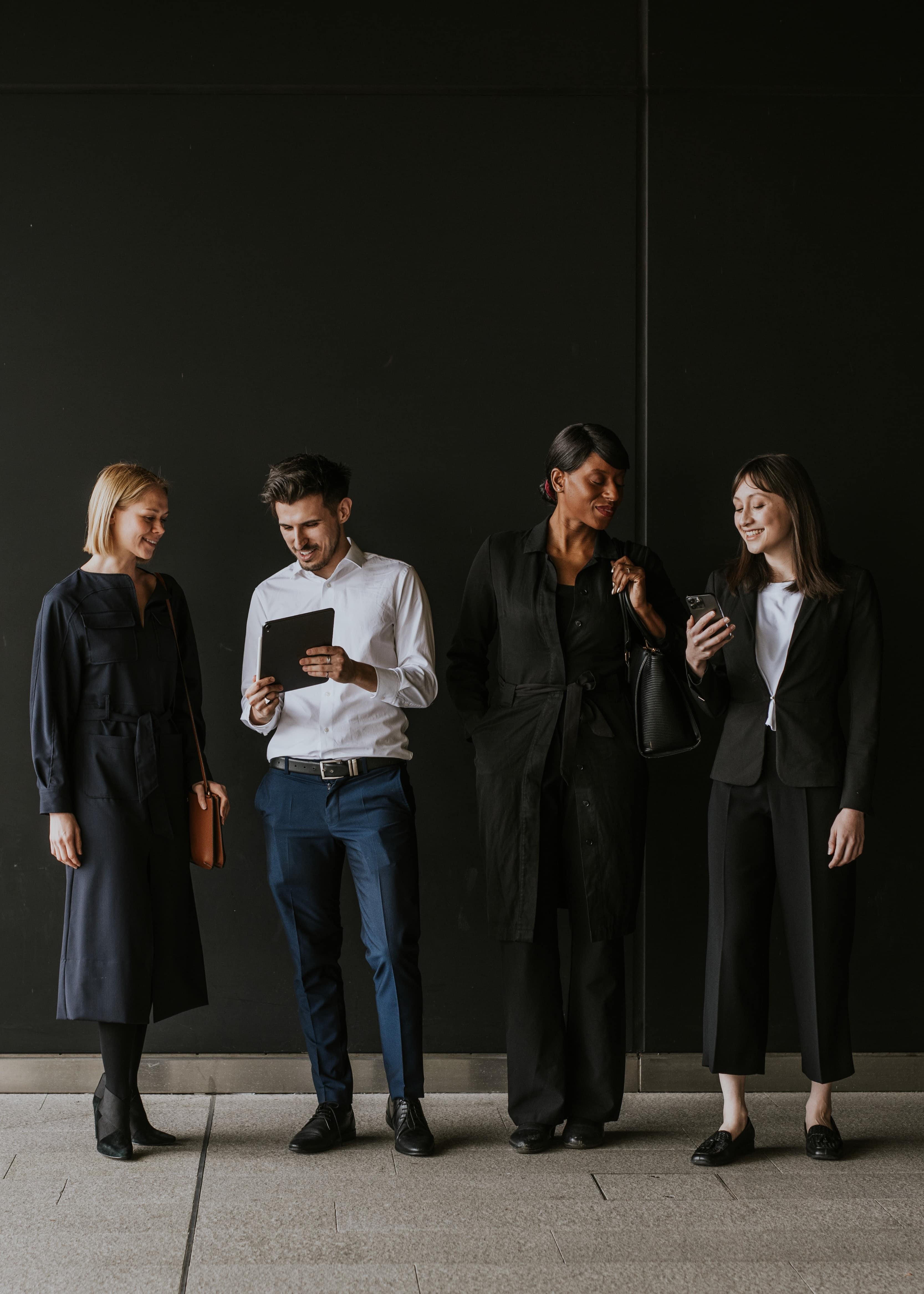 Four professionals in dark business attire stand against a black wall; one man reads a tablet while two women look on, one using a smartphone.