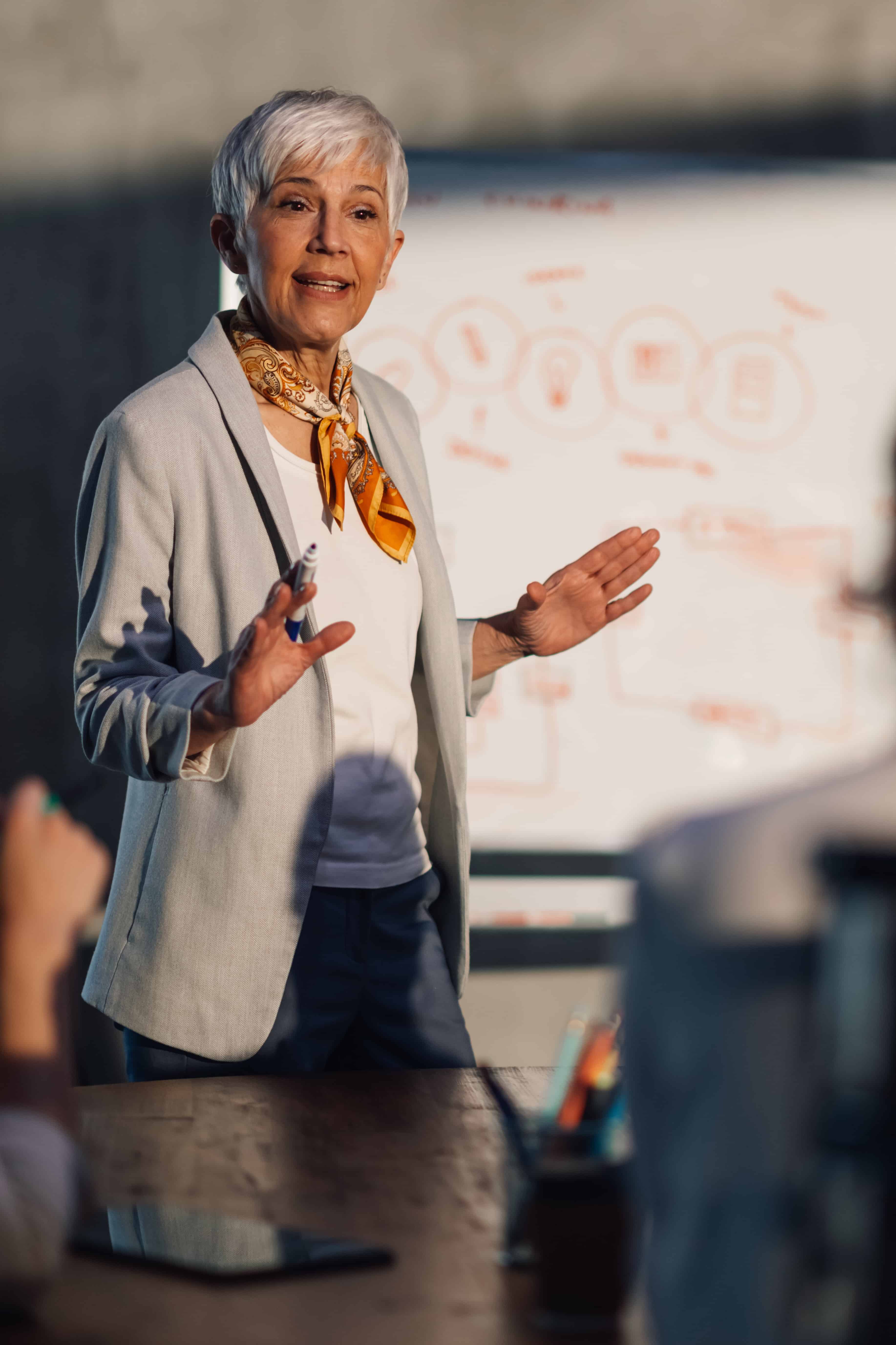 Older woman presenter with short white hair, gray blazer and orange scarf addressing an audience.