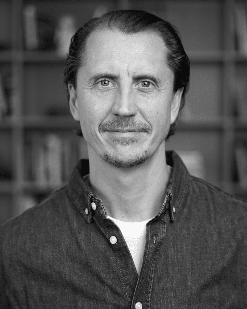 Black-and-white close-up portrait of a man with dark hair and a light moustache wearing a denim shirt, standing in front of a blurred bookshelf backdrop.