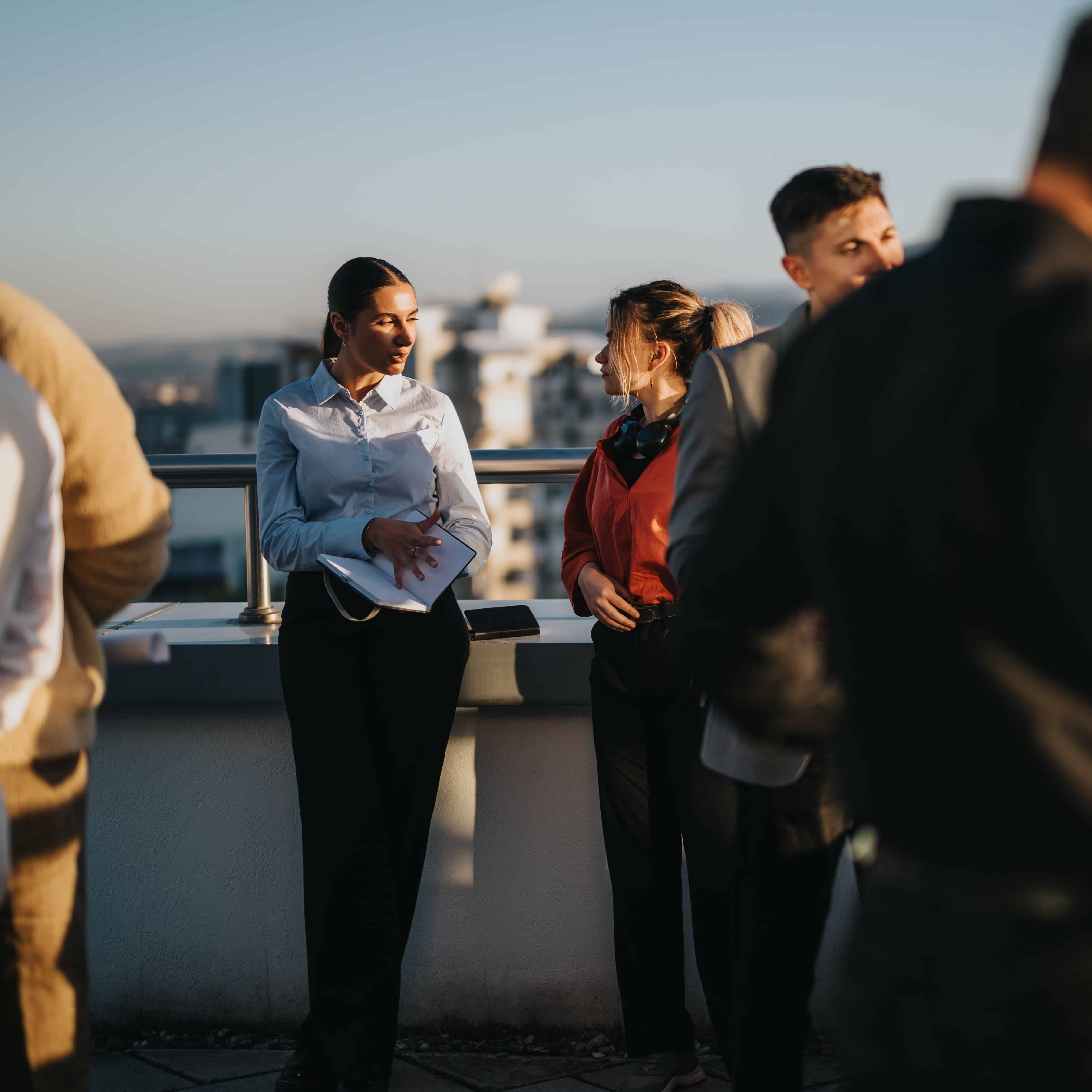 Group of people standing and talking on a rooftop with city buildings in the background during sunset sun.