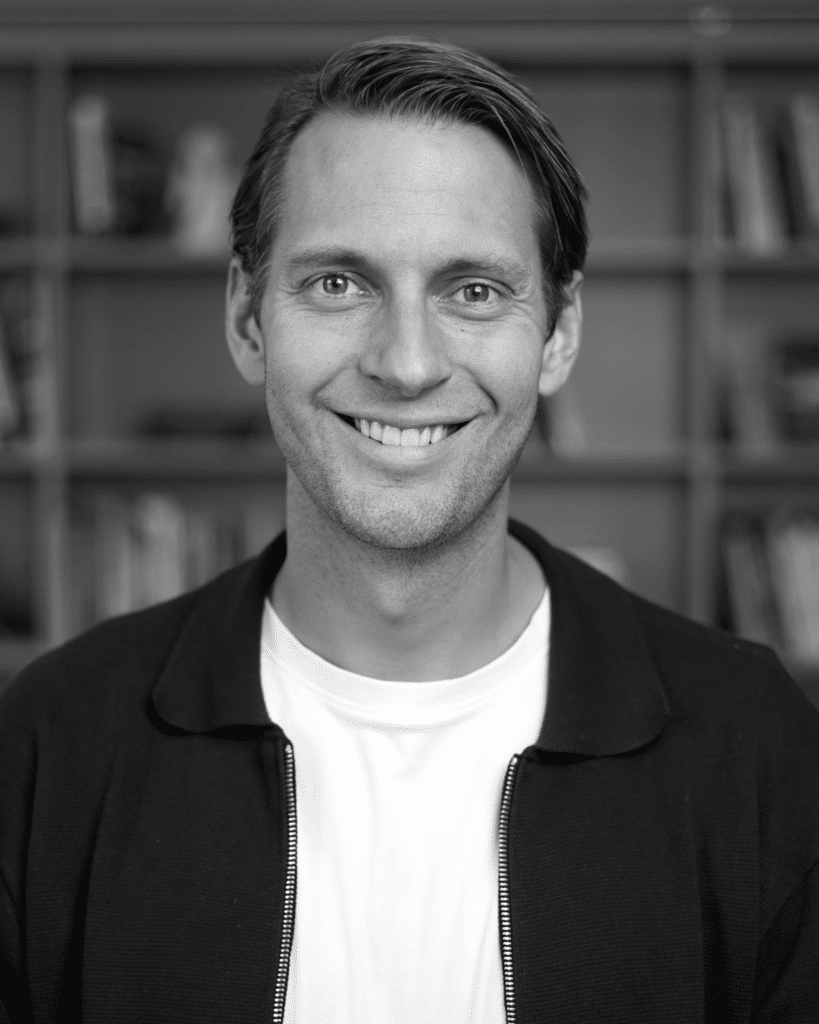 Close-up of a smiling man with short hair, wearing a white shirt and black jacket, blurred bookshelves in the background.
